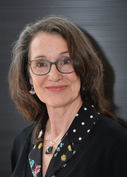 Portrait de l'artiste peintre Christine Bouchard, devant un fond gris strié, portant des lunettes et un chemisier fleuri.